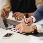 Dos personas sentadas ante un escritorio con un ordenador portátil discuten sobre la ley de la segunda oportunidad. Una escribe en un portapapeles mientras la otra gesticula. Un smartphone y una pequeña planta descansan sobre la mesa, con la atención puesta en sus manos y materiales de trabajo.