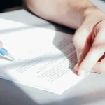 Primer plano de las manos de una persona que sostiene un bolígrafo y escribe en un documento impreso sobre la ley de la segunda oportunidad en un escritorio, con la luz del sol proyectando sombras sobre el papel.